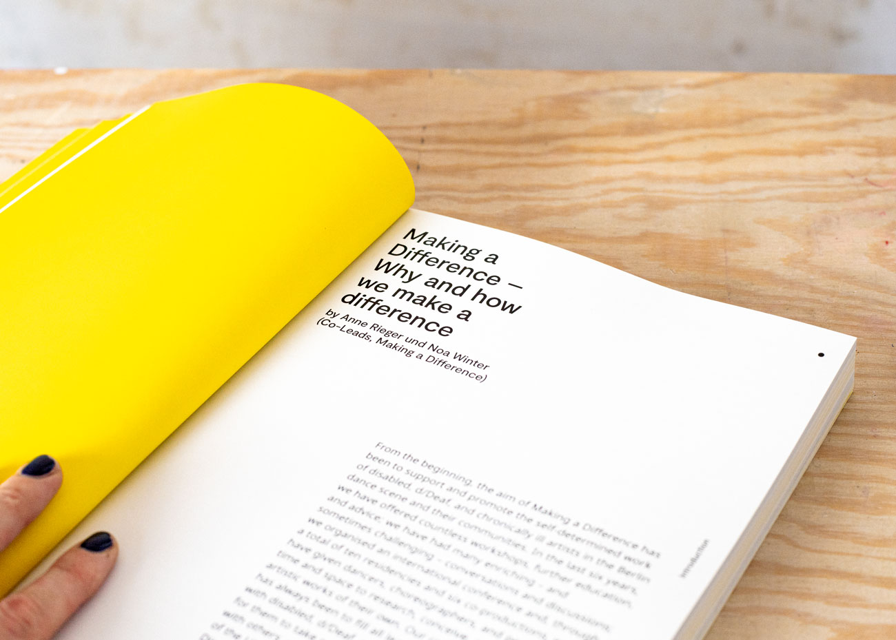 Detailaufnahme einer Doppelseite des Buchs auf hellem Holz-Untergrund. Die linke Seite ist komplett Gelb. Darauf liegt eine Hand mit dunkelblau lackierten Nägeln. Auf der rechten Seite befinden sich in schwarz auf weiß die Überschrift, ein Autor*innen-Hinweis und Text.