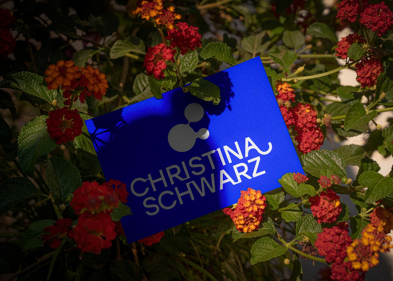 Die bleue Visitenkarte mit dem weißen Logo liegt in einem grünen Strauch, gesäumt von roten und orangenen Blumen. Der Strauch wirft starke Schatten auf Teile der Visitankarte.
