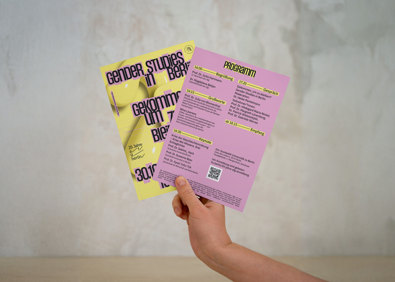 Hand hält zwei Flyer, einer gelb mit schwarzem Text, einer rosa mit Programmtext und QR-Code, vor unscharfem Hintergrund