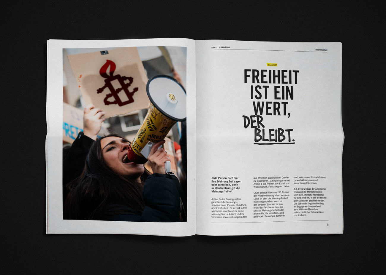 Die Doppelseite einer Zeitung zeigt links ein Foto einer Person mit einem Megafon und einem Schild mit dem Amnesty-International-Logo in Rot und Schwarz. Rechts befindet sich eine Überschrift in großer, schwarzer Schrift: »FREIHEIT IST EIN WERT, DER BLEIBT.« Darunter steht ein mehrspaltiger Fließtext in kleinerer Schrift. Das Layout ist klar und übersichtlich, mit viel Weißraum und einer modernen, serifenlosen Schriftart.