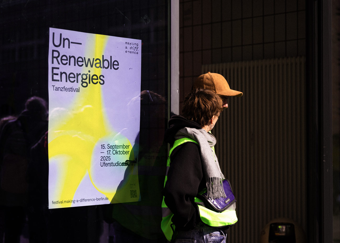 Das Plakat zeigt den Titel Un-Renewable Energies in großen schwarzen Buchstaben auf weißem Hintergrund mit einem gelben Farbverlauf. Darunter steht Tanzfestival und die Daten 15. September – 17. Oktober 2025 Uferstudios. Oben rechts befindet sich der Schriftzug making a difference. Unten links ist die Webseite festival.making-a-difference-berlin.de angegeben. Das Plakat ist an einer dunklen Glasfläche angebracht, vor der zwei Personen mit Jacken und Schals stehen, von denen eine eine beige Kappe trägt.