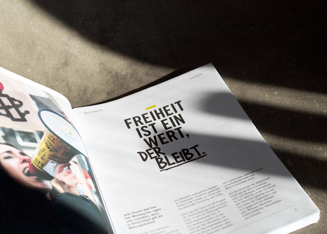 Die Doppelseite einer geöffneten Zeitung. Auf der rechten Seite eine große, mittig platzierte Überschrift in schwarzer Schrift: 'FREIHEIT IST EIN WERT, DER BLEIBT.' Darunter befindet sich ein mehrspaltiger Fließtext in kleinerer Schrift. Die linke Seite zeigt ein Foto einer Person mit einem gelben Megafon in der Hand. Die Seite liegt auf einem Beton-Untergrund, und ein Schatten fällt diagonal über die rechte Seite der Zeitung.