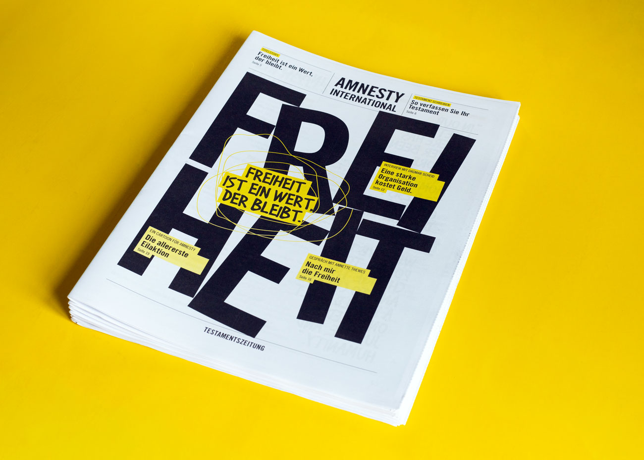 Ein auf einem gelben Untergrund liegender Stapel Zeitungen mit dem Titel 'FREIHEIT' in großen, schwarzen Großbuchstaben. Auf dem Titelblatt steht oben 'AMNESTY INTERNATIONAL' in schwarzer Schrift. In der Mitte des Titels befindet sich der gelb hinterlegte schwarze Text 'FREIHEIT IST EIN WERT, DER BLEIBT.' Weitere gelbe Textfelder mit schwarzer Schrift sind auf dem Titelblatt verteilt, darunter.