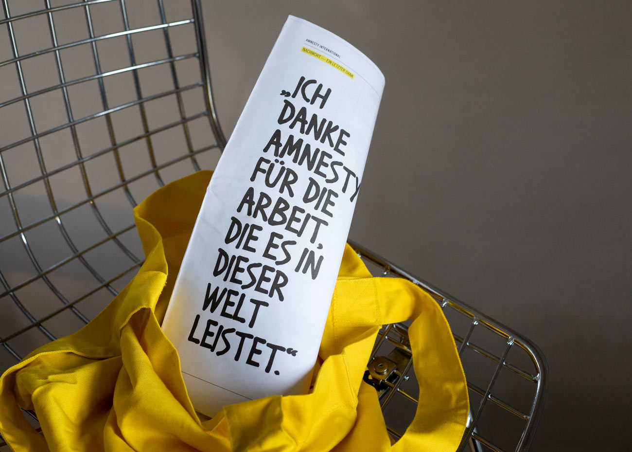 Eine zusammengerollte Zeitung mit handschriftlich wirkendem, schwarzem Text steht aufrecht in einer leuchtend gelben Stofftasche, die auf einem silbernen Stuhl liegt. Der Text auf dem Papier lautet: '„ICH DANKE AMNESTY FÜR DIE ARBEIT, DIE ES IN DIESER WELT LEISTET.“'. Der Hintergrund ist einfarbig beigebraun und unscharf, wodurch der Fokus auf dem Papier und der Tasche liegt.