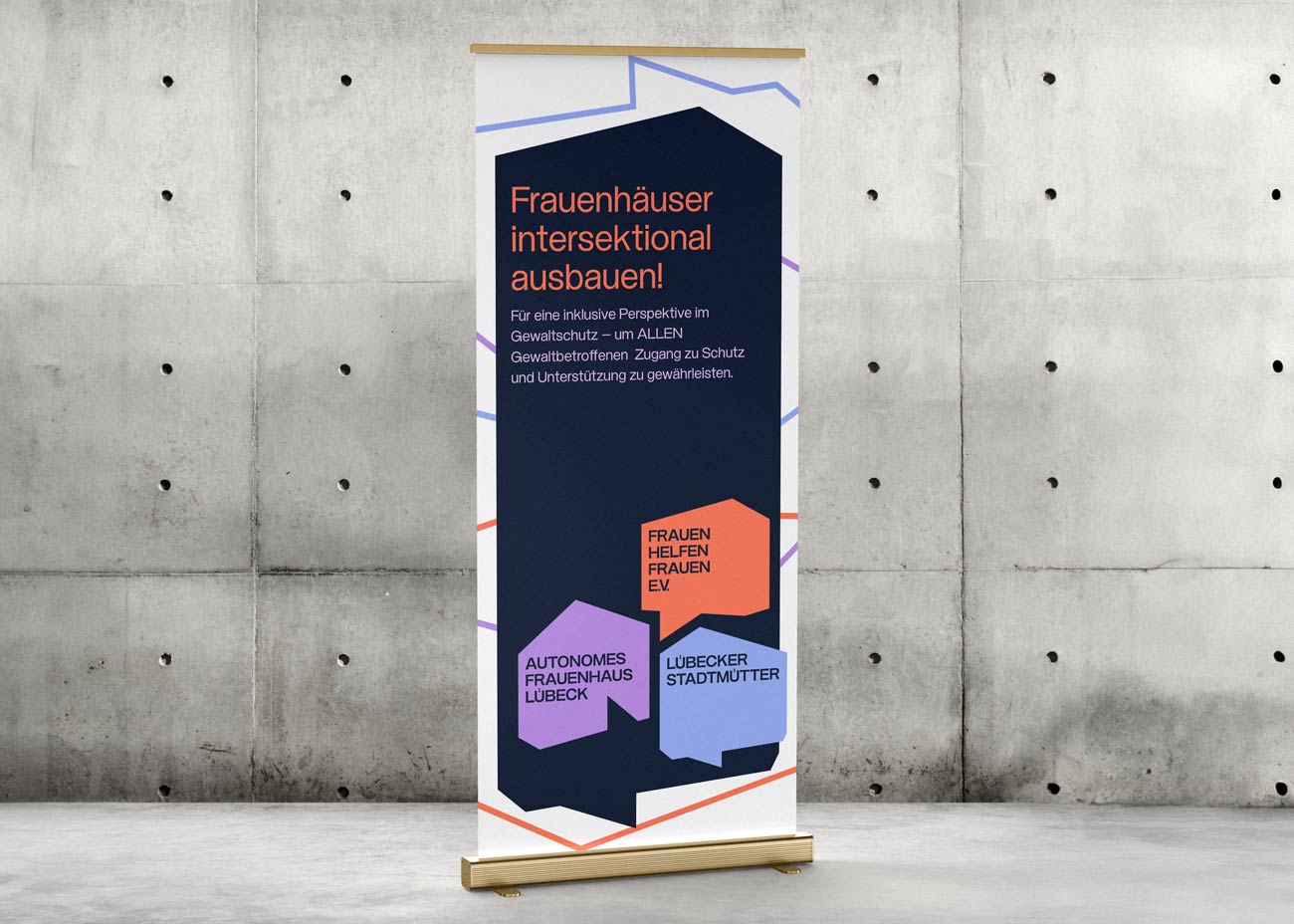 Ein Roll-up-Banner vor einer grauen Betonwand. Das Banner zeigt ein modernes, geometrisches Design mit dunkelblauem Hintergrund und farbigen polygonalen Formen in Orange, Lila und Hellblau. Oben steht in orangefarbener Schrift der Text 'Frauenhäuser intersektional ausbauen!'. Darunter folgt ein erklärender Text in weißer Schrift. Unten sind die drei bunten Logos der Bereiche kombiniert: 'FRAUEN HELFEN FRAUEN EV' in Orange, 'AUTONOMES FRAUENHAUS LÜBECK' in Lila und 'LÜBECKER STADTMÜTTER' in Hellblau. 