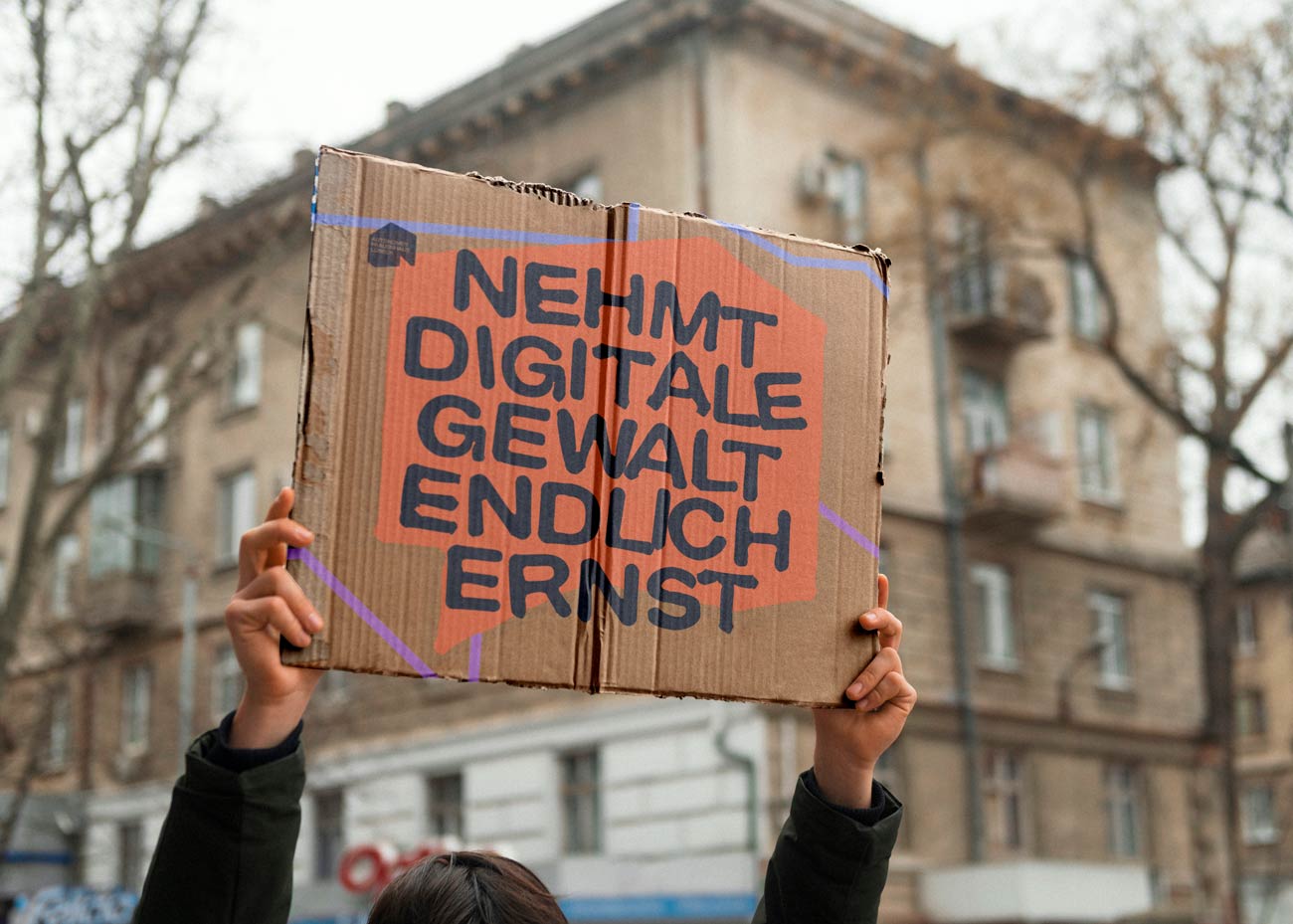 Zwei Hände halten ein rechteckiges Demoschild mit schwarzer Schrift und einer orangefarbenen große Sprechblase hoch. Auf dem Schild steht in Großbuchstaben: 'NEHMT DIGITALE GEWALT ENDLICH ERNST'. Im Hintergrund ist unscharf ein mehrstöckiges Gebäude mit mehreren Fenstern zu sehen.