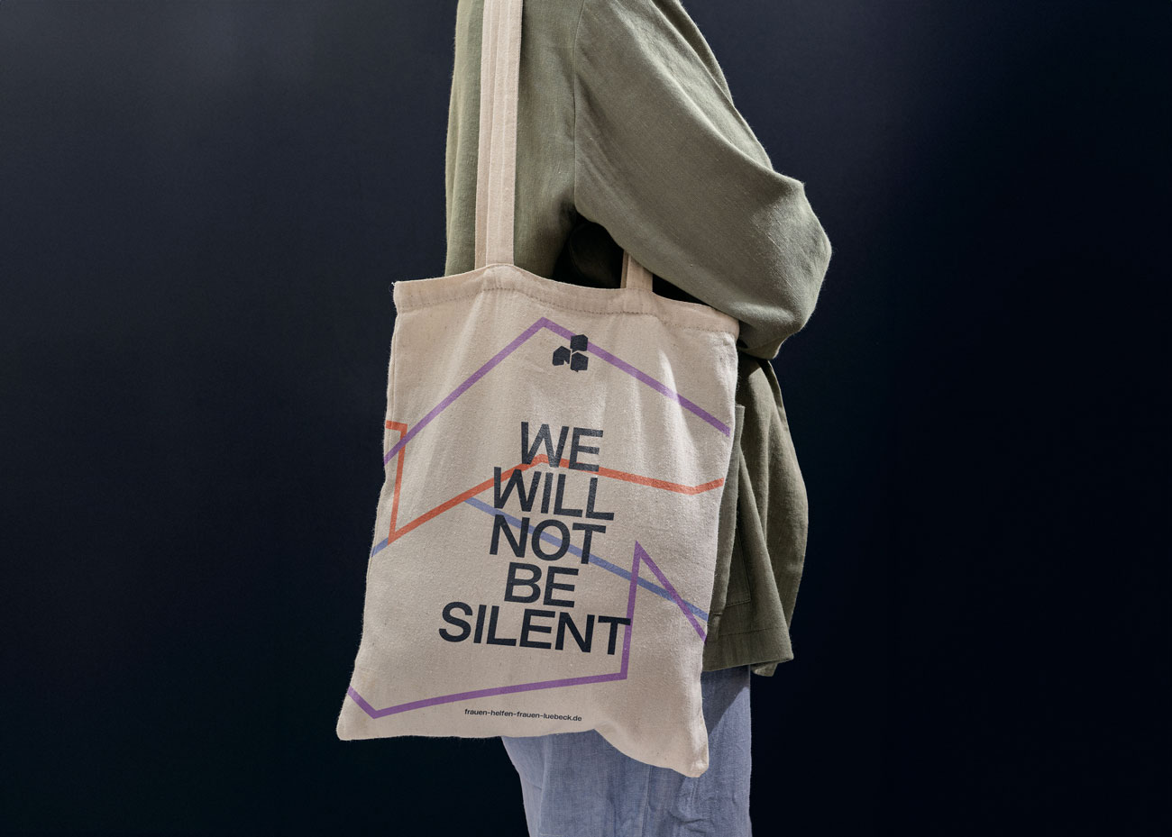 Eine Hand hält eine orangefarbene Stofftasche mit langen Henkeln vor einer grauen Betonwand. Die Tasche zeigt einen lilafarbenen geometrischen Farbblock im unteren Bereich. Darauf steht in großen schwarzen Großbuchstaben der Text 'WE WILL NOT BE SILENT'. Darüber in klein die drei dunkelblauen Logos des Vereins. Unten die Webadresse.