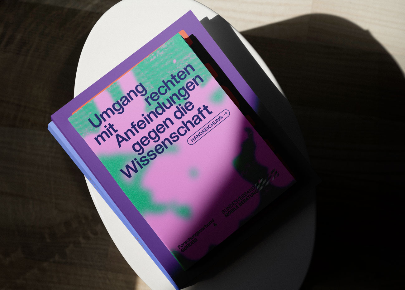 Mehrere Bücher liegen übereinander auf einem Tisch. Der Titel des oberen Buches ist in dunkler Schrift auf dem Umschlag zu lesen: 'Umgang mit rechten Anfeindungen gegen die Wissenschaft'. Darunter steht Handreichung in einem kleinen, abgerundeten Rechteck. Der Umschlag zeigt ein abstraktes Muster in Grün- und Pink. Das Buch liegt in einem Bereich mit Licht- und Schattenkontrasten, wobei ein Teil des Buches im Schatten liegt.