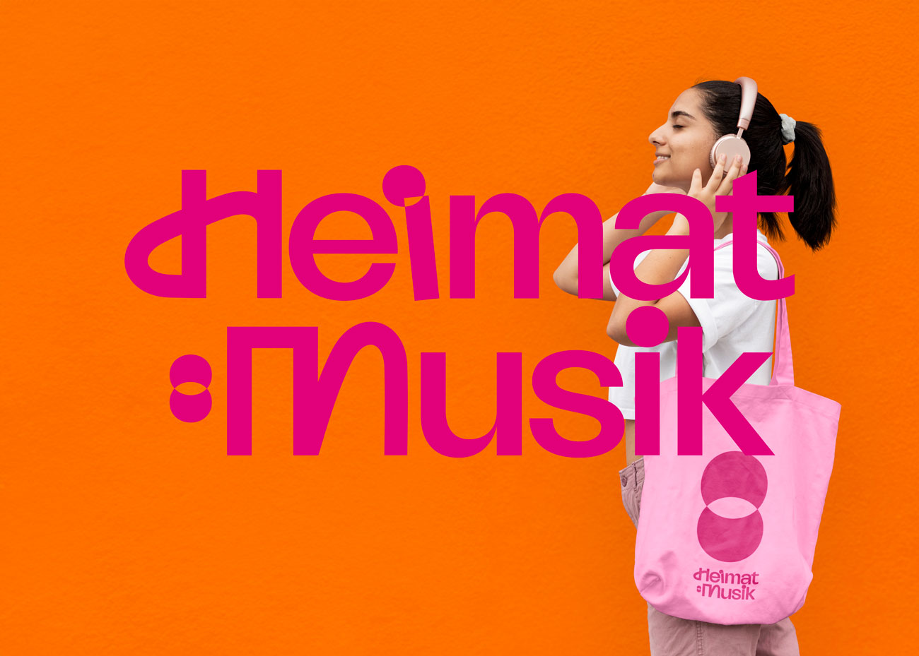 Vor einem einfarbigen, leuchtend orangen Hintergrund steht eine Person im Profil, die nach links blickt. Sie trägt ein weißes T-Shirt und eine hellrosa Stofftasche mit der pinkfarbenen Grafik und dem Logo 'heimat:musik'. Die Person hat dunkle Haare, die zu einem Zopf gebunden sind, und hält Kopfhörer an ihr Ohr. Auf dem Foto ist das Logo von 'Heimat:Musik' in einer modernen, verspielten pinkfarbenen Schrift mittig im Bild platziert.