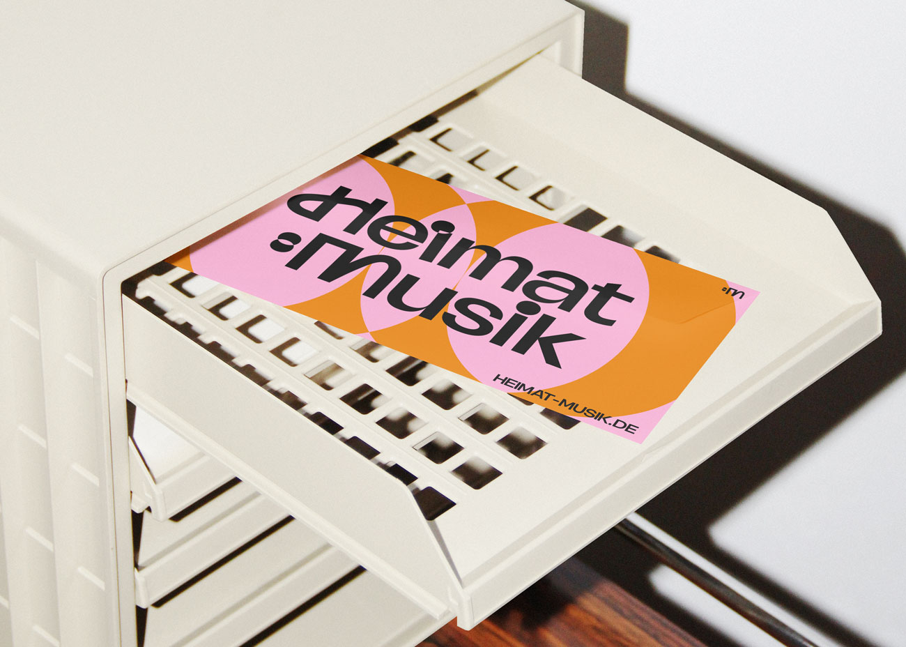 Auf einem weißen Aktenregal liegt eine Visitenkarte. Der Hintergrund der Karte ist grafisch gestaltet und besteht aus zwei Farben: ein orangefarbener Hintergrund und darüberliegende, sich überlappende rosa Kreise. Darauf liegt in schwarzer Schrift das Logo von ‚Heimt:Musik'. Unten rechts auf der Karte befindet sich in kleinerer schwarzer Schrift die Webadresse 'HEIMAT-MUSIK.DE'.