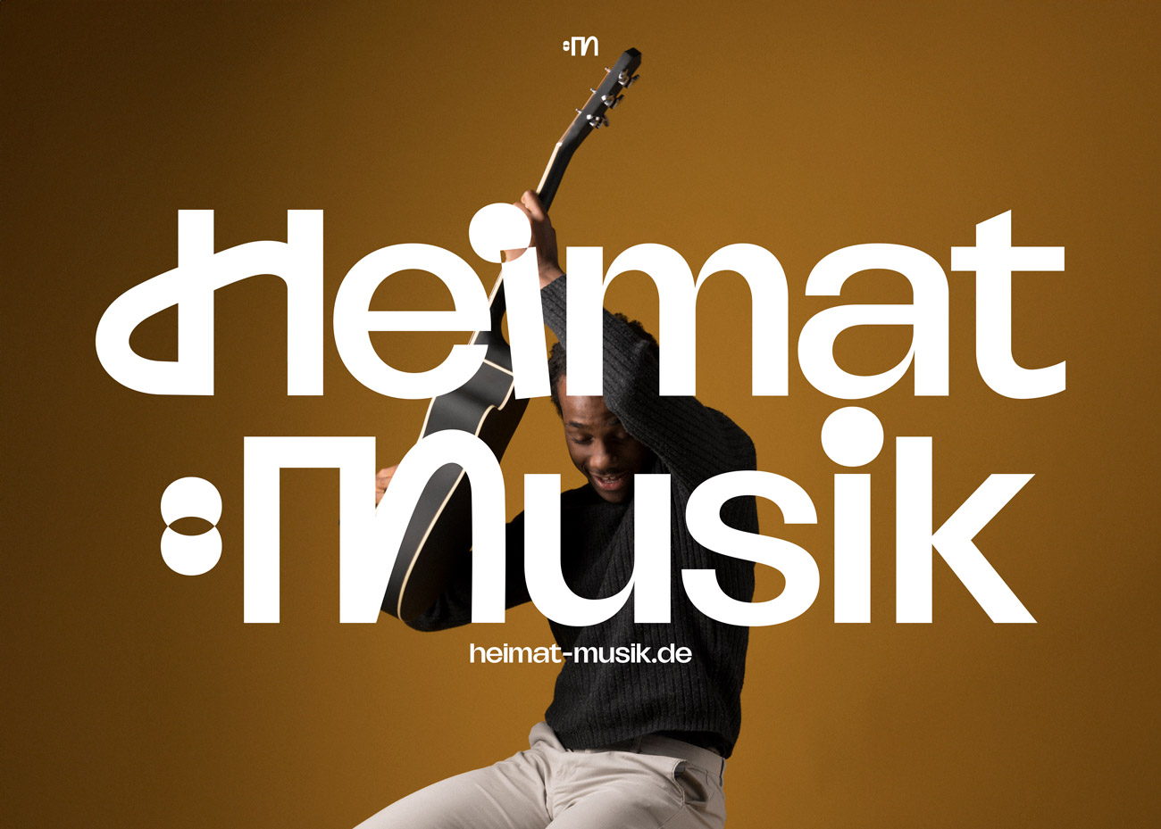 Eine Person in einem schwarzen Pullover und heller Hose hält eine Gitarre über den Kopf und lacht dabei. Der Hintergrund ist einfarbig in einem warmen Braunton gehalten. Über der Person steht in großen, weißen Buchstaben das Logo ‚Heimat:Musik'. Unter dem Schriftzug ist in  weißer Schrift die Webadresse 'heimat-musik.de' zu sehen.