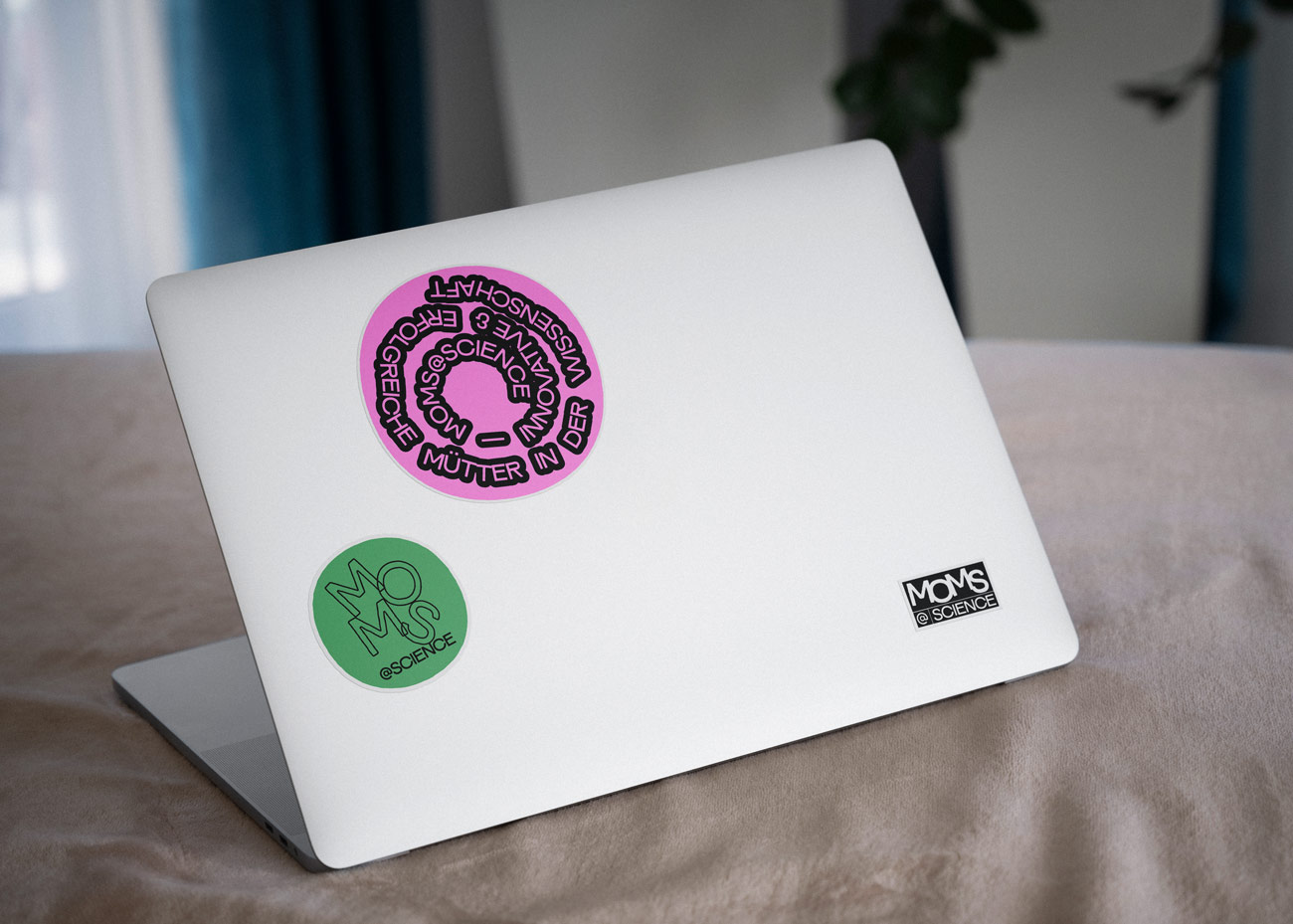 Auf der Rückseite eines silberfarbenen Laptops befinden sich drei Aufkleber. Ein großer, runder, pinkfarbener Aufkleber zeigt in schwarzer Schrift den Text 'MÜTTER IN DER WISSENSCHAFT' kreisförmig um das Wort 'MOMS' in der Mitte. Ein kleinerer, runder, grüner Aufkleber trägt in schwarzer Schrift das Wort 'MOMS' und darunter '@SCIENCE'. Ein rechteckiger, schwarzer Aufkleber mit weißer Schrift zeigt ebenfalls den Schriftzug 'MOMS @SCIENCE'.