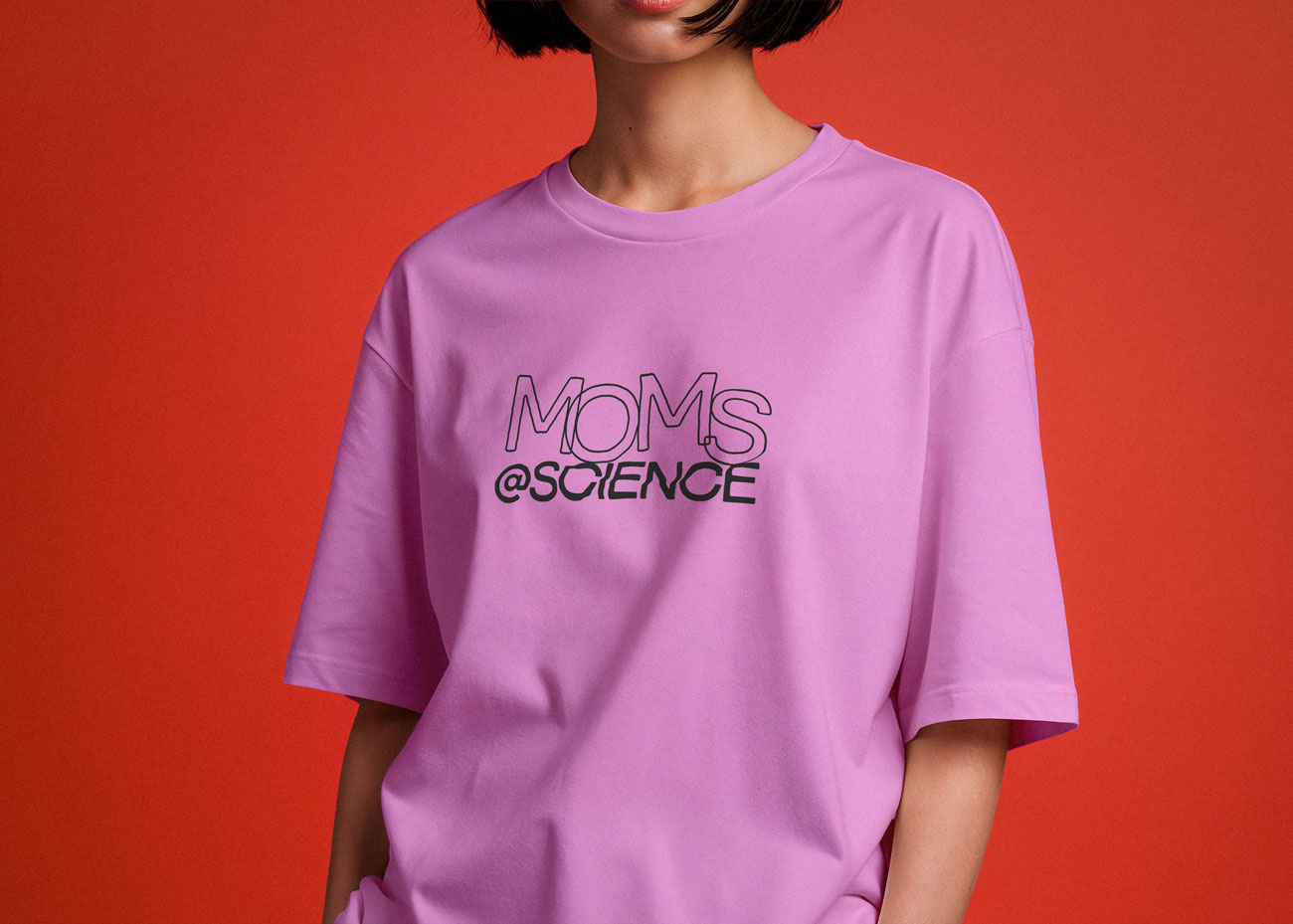 Eine Person trägt ein locker sitzendes, rosafarbenes T-Shirt vor einem rötlichen Hintergrund. Auf dem T-Shirt steht in schwarzer Schrift 'MOMS @SCIENCE'. Der obere Teil des Gesichts der Person ist nicht sichtbar, nur der Mund und das Kinn sind zu sehen. Die Person hat kurze, dunkle Haare.