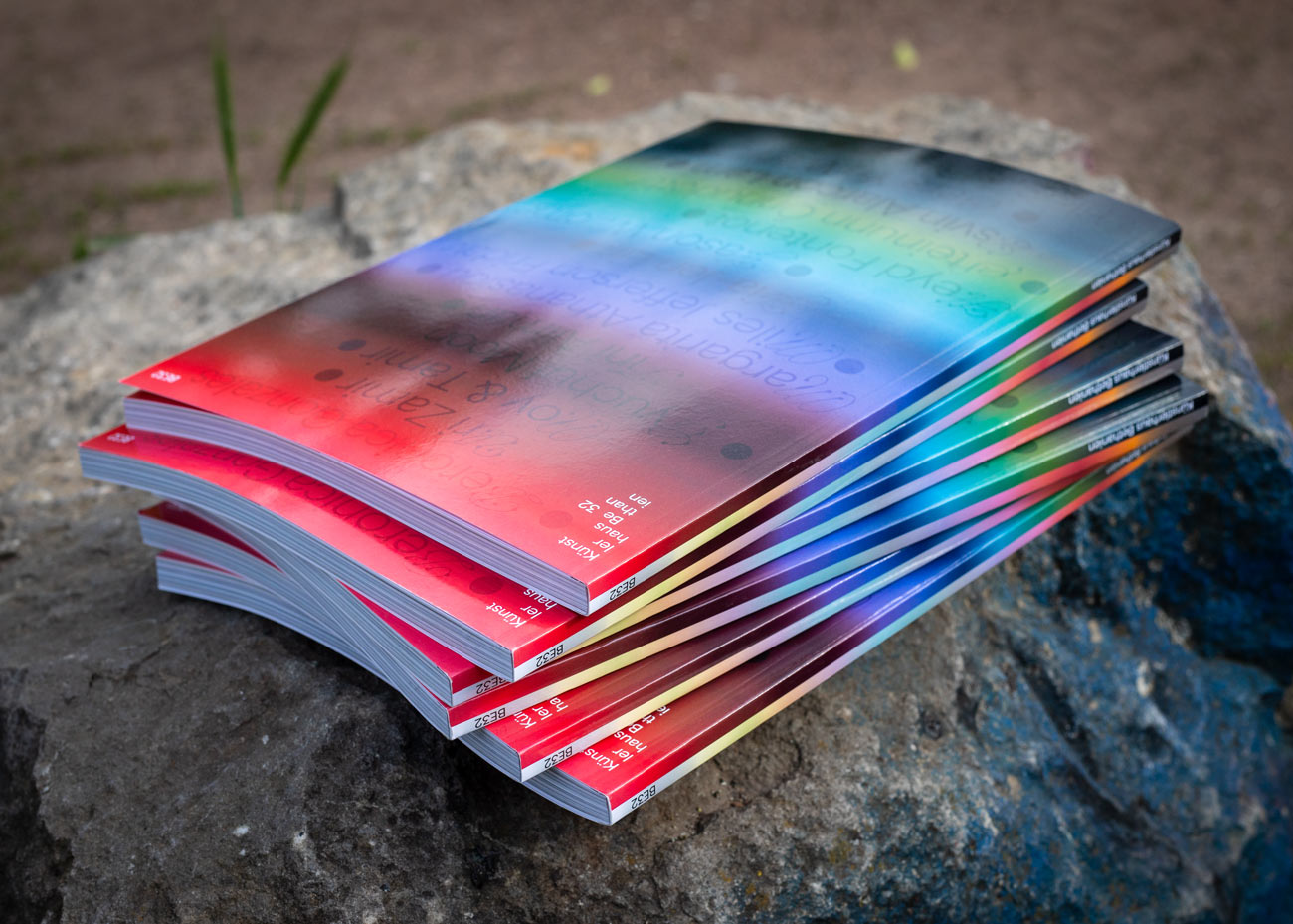 Mehrere Ausgaben des Magazins liegen gestapelt auf einem großen Stein. Die Covergestaltung zeigt einen auffälligen Farbverlauf von Rot über Blau zu Grün, der sich über die gesamte Titelseite zieht. Die Magazine sind leicht versetzt übereinandergelegt, sodass die farbigen Kanten sichtbar werden. Im oberen Bereich des Covers ist eine weißer Schriftzug erkennbar. Der Hintergrund ist unscharf und zeigt einen sandigen Boden.