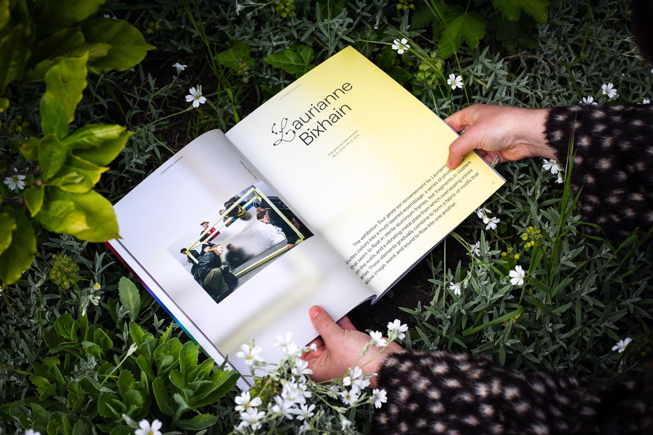 Eine Person mit einer braun-weiß gepunkteten Jacke hält das Magazin über einer Blumenwiese. Die Doppelseite zeigt links das Bild einer Ausstellung und rechts auf hellgelben Grund in großer Schrift mit einer schnörkeligen Versalie den Namen Laurianne Bixhain. Darunter realtiv groß gesetzter Text.