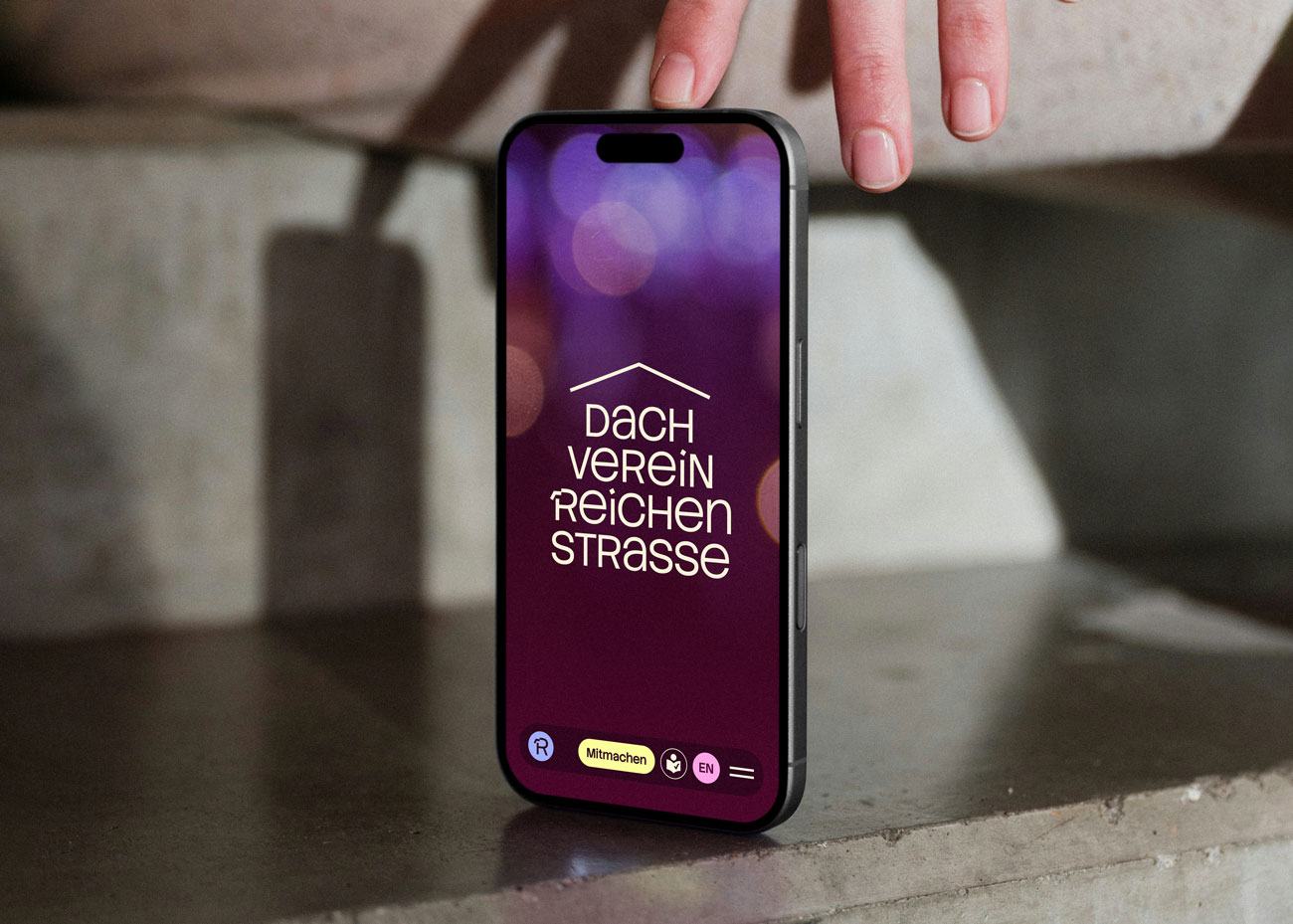 Ein Smartphone befindet sich zwischen einem Betonboden und einer Hand. Auf dem Display ist die Startseite des Dachverein Reichenstraße zu sehen, im Fokus sind das Logodesign und die Navigationsleiste mit den barrierefreien Farben und Icons zu Sprache, leichte Sprache und dem Menu.