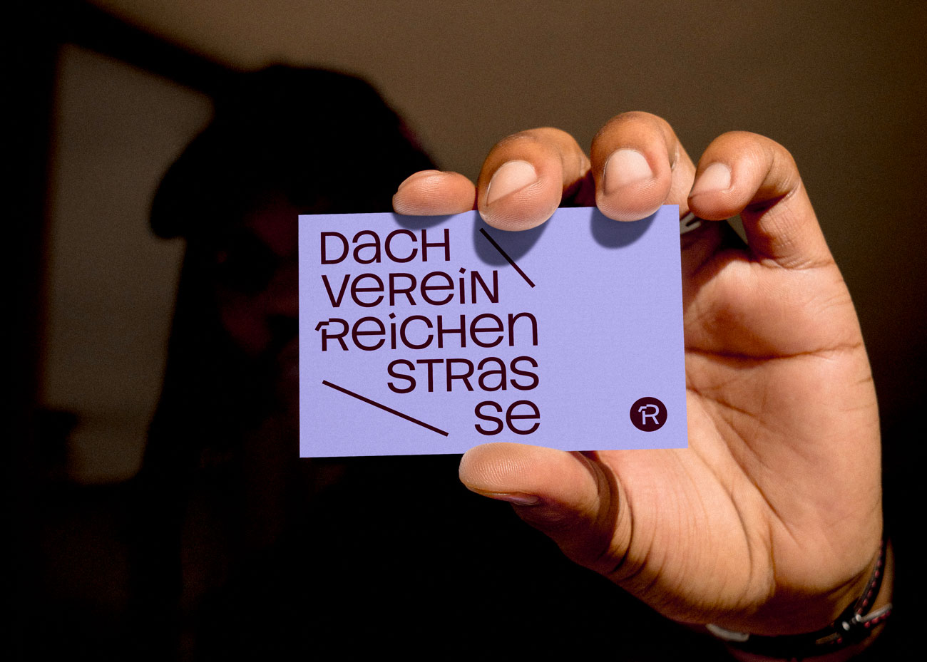 Eine Person hälte eine lila Visitenkarte mit dem Typografischen Logo des Dachverein Reichenstraße. Es besteht aus locker gesetzten Buchstaben und Linien. Daneben ein kleines Bildzeichen in dunklem Rot, bestehend aus dem R in einem Kreis.
